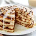Fluffy cinnamon roll pancakes drizzled with icing atop a breakfast plate.